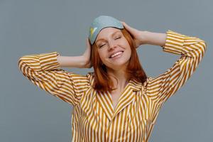 une jeune femme rousse heureuse a assez dormi, garde les mains sur la tête, se tient les yeux fermés, porte des vêtements confortables et un bandeau sur les yeux, isolée sur fond gris, se repose bien. notion de temps de sommeil photo