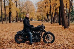 photo extérieure d'un motard à barbe épaisse, porte des gants de protection, un manteau noir et un jean, pose sur une moto dans un magnifique parc aux feuilles d'oranger, tient un casque, s'arrête pour se reposer. mode de vie urbain