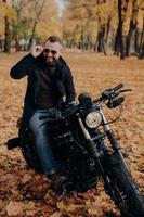 image d'un motocycliste masculin joyeux sourit joyeusement, garde la main sur les nuances, s'assoit sur un vélo rapide, s'amuse en plein air, aime voyager en moto, passe du temps libre dans le parc d'automne. notion de voyage. photo