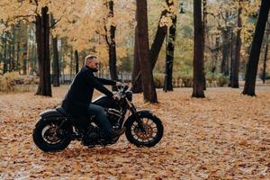 image extérieure d'un motocycliste masculin pose sur une moto rapide, porte des nuances, un manteau noir, aime rouler dans le parc d'automne, respire l'air frais, admire la nature pendant la journée ensoleillée. homme barbu actif à l'extérieur photo