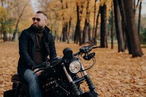 photo extérieure d'un motard pensif qui garde le regard de côté, a un regard pensif, porte des lunettes de soleil, un pull, une veste noire et un jean, pose sur une moto en plein air dans le parc d'automne. liberté, style de vie, passe-temps