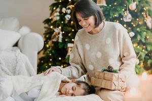 j'ai un petit secret pour toi. belle mère et fille célèbrent le nouvel an à la maison. maman pose près d'une fille endormie avec des cadeaux contre l'arbre du nouvel an décoré. concept de joyeuses vacances d'hiver photo