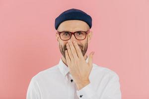 un homme positif non rasé couvre la bouche avec la paume, rit positivement, porte des lunettes, entend une blague amusante, des modèles sur fond rose. un designer masculin heureux reçoit de bonnes nouvelles. notion d'émotions photo