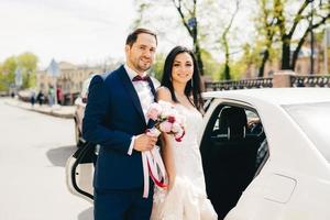 un joli couple marié se tient l'un à côté de l'autre près de la voiture, va au restaurant pour célébrer son mariage, faire la fête, inviter des amis et des parents. les jeunes mariés posent en plein air après l'enregistrement du mariage photo