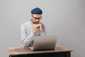 un bourreau de travail masculin sérieux est assis au bureau avec un appareil électronique moderne, regarde une vidéo, prépare un rapport financier, se concentre sur un ordinateur portable, porte une chemise blanche, des lunettes optiques, des modèles à l'intérieur photo