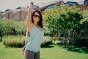 belle jeune femme heureuse fait des exercices d'armes avec des supports de gomme de remise en forme en plein air a un corps athlétique porte des lunettes de soleil aime la journée d'été a une formation régulière. la sportive utilise un extenseur. notion de sport photo