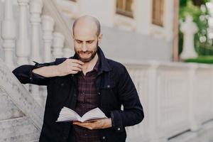 beau mâle chauve intelligent tient un livre, lit un roman intéressant avec une expression sérieuse, se tient à l'extérieur contre un ancien bâtiment blanc, aime lire. concept de personnes, passe-temps, style de vie et loisirs photo