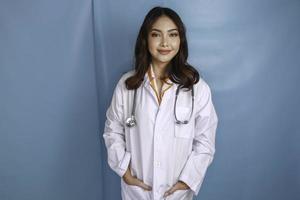 une femme médecin asiatique détendue et confiante debout avec les mains dans les poches de sa blouse de laboratoire souriant tranquillement à la caméra sur fond bleu avec espace de copie photo