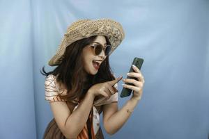 jeune femme heureuse asiatique pointant et souriant vers son smartphone isolé par un fond bleu photo