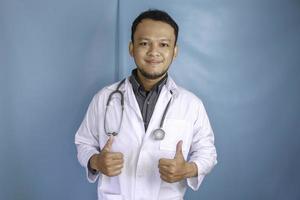 portrait d'un jeune médecin asiatique, un professionnel de la santé sourit et montre le pouce levé ou le signe ok isolé sur fond bleu photo