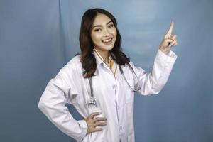 portrait d'une jeune femme médecin asiatique, un professionnel de la santé sourit et pointe vers le haut sur un espace de copie isolé sur fond bleu photo
