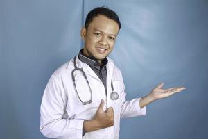 heureux jeune homme asiatique médecin, professionnel de la santé sourit et pointe vers un espace de copie isolé sur fond bleu photo