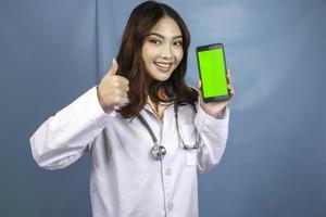 une jeune femme médecin asiatique pointe et montre un écran vert ou un espace de copie sur son smartphone. montrant les pouces vers le haut ou le signe ok. photo