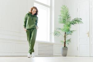 photo pleine longueur d'une jeune femme active et heureuse avec un corps mince, garde les mains dans les poches du survêtement, concentrée vers le bas, prête à faire des exercices cardio, a un look déterminé, pilates faisant de l'exercice à l'intérieur