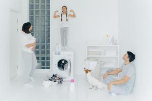 photo d'une écolière heureuse pose sur le dessus de la machine à laver, montre les muscles, lève les bras, prête à aider les parents à laver ou à laver. femme et homme posent dans la salle de lavage avec chien et petit enfant