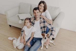 vue de dessus des membres heureux de la famille posent dans une pièce spacieuse près d'un canapé confortable, le chien préféré pose à proximité, profitez du temps libre assis sur le sol, souriez positivement. père, mère et enfant passent du temps à la maison photo