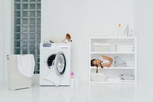 une petite fille fatiguée a travaillé dans la maison pour aider les parents, dort sur une étagère près du linge plié, du fer et des contenants de lessive, jack russell terrier sur la machine à laver. enfants, concept de tâches ménagères photo