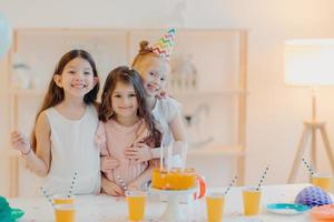 photo horizontale de trois amis heureux s'embrassent et ont des expressions amusantes, posent près de la table de fête avec un gâteau à l'intérieur. trois filles fêtent leur anniversaire ensemble, s'amusent, viennent faire la fête