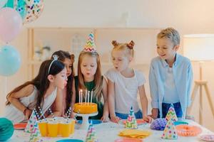 les petits enfants célèbrent la fête d'anniversaire, soufflent des bougies sur le gâteau, se rassemblent à une table de fête, ont de la bonne humeur, passent du temps ensemble, font des vœux, portent des chapeaux de fête, posent à l'intérieur avec des ballons gonflés