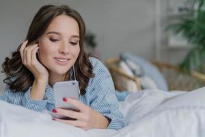 belle femme écoute de la musique, passe un appel vidéo, se trouve dans un lit confortable le matin, habillée avec désinvolture, a un sourire charmant, pose dans la chambre. concept de personnes, de détente et de technologies modernes photo