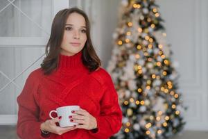 photo intérieure d'une jeune femme réfléchie vêtue de vêtements d'hiver chauds, apprécie le café chaud ou le cappuccino, regarde pensivement de côté, se tient près de l'arbre de noël décoré, a une manucure rouge. notion de confort