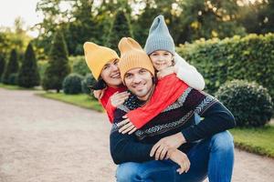 bel homme a de bonnes relations avec sa femme et sa petite fille qui l'embrassent de dos, ont des sourires agréables. une jeune famille sympathique en bonnets et chandails tricotés se tient contre des arbres verts photo