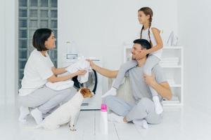 famille, ménage, concept de lavage. un père souriant donne un tour de ferroutage à une petite fille donne à sa femme un t-shirt blanc à laver est assis sur le sol de la salle de lavage en pose de lotus, animal domestique près de la machine à laver photo