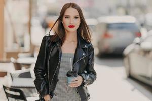 photo en plein air d'un modèle féminin attrayant du millénaire avec du maquillage, vêtu d'une veste en cuir élégante, boit du café dans une tasse jetable, se promène dans la ville, profite de ses loisirs. personnes, concept de boisson