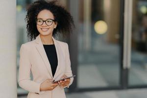 une femme entrepreneure réussie et positive aux cheveux afro tient une tablette numérique, se tient à l'extérieur près d'un immeuble de bureaux, porte des vêtements formels, regarde ailleurs, attend que son collègue dîne pendant la pause photo