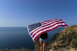 vue arrière d'une jeune femme tenant un drapeau américain agitant sur la côte contre la mer ensoleillée. photo