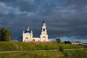 une vieille église orthodoxe et un village au bord de la rivière. coucher de soleil et nuages noirs. photo