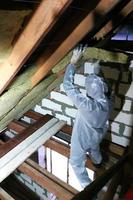 un homme dans une combinaison de protection met de la laine minérale entre la poutre du toit de la maison pour son réchauffement du froid photo