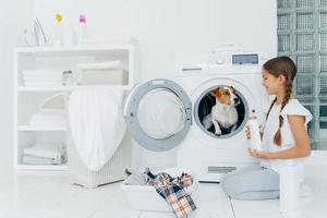 positive petite fille s'amuse avec jack russel terrier, pose sur le sol près de la machine à laver dans la buanderie, tient de la poudre liquide, occupée avec les travaux ménagers, a une expression heureuse. intérieur cosy dans la salle de bain photo