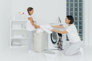 une mère heureuse charge des vêtements dans la machine à laver, une petite fille aide, donne du linge blanc du panier, fait le ménage ensemble, vêtue de vêtements décontractés. fille offre de l'aide à maman, nettoie les vêtements photo