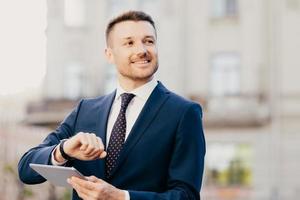 séduisant homme d'affaires en costume noir élégant, regarde la montre-bracelet, tient une tablette moderne, envoie des messages avec des amis et des collègues sur les réseaux sociaux, se réunit trop tôt. notion de carrière photo