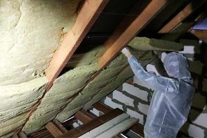 un homme dans une combinaison de protection met de la laine minérale entre la poutre du toit de la maison pour son réchauffement du froid photo