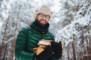 beau mâle barbu en lunettes et chapeau chaud avec anorak, tient du bois de chauffage, pose contre des arbres recouverts de neige blanche étincelante, passe du temps en forêt. tir extérieur d'un homme heureux repose dans la forêt photo