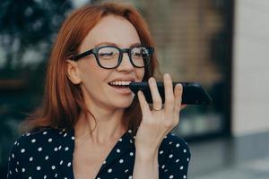 femme d'affaires souriante aux cheveux roux utilisant un assistant vocal numérique virtuel tout en se tenant à l'extérieur