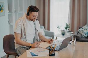 photo en intérieur d'un homme heureux prend des notes tout en écoutant des cours en ligne étudie à distance depuis son domicile est assis au bureau avec des papiers d'ordinateur portable modernes porte des écouteurs a une conférence Web ou une réunion virtuelle