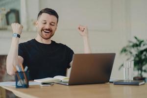 un homme barbu joyeux et excité avec une expression joyeuse serre les poings célèbre le succès habillé avec désinvolture pose au bureau devant un ordinateur portable ouvert satisfait des résultats du travail. oui super boulot photo