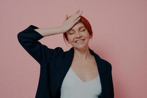 belle femme rousse insouciante en riant isolée sur fond de studio rose en gardant la main sur le front photo