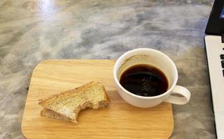 tasse de café noir restant avec un sandwich au fromage au jambon mordu sur une planche de bois et une table en béton gris photo