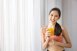 femme asiatique en bonne santé buvant un verre de jus d'orange pour se rafraîchir tout en portant des vêtements de sport pour la nutrition et des suppléments de vitamine c à la fenêtre avec espace de copie photo