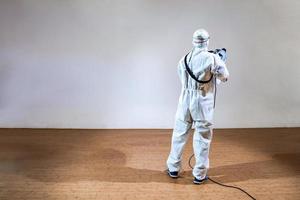 un homme technique professionnel en combinaison de prévention pulvérise une solution de stérilisation par une machine de pulvérisation électrique sur un sol en bois et un fond blanc avec une lumière de studio. photo