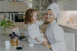 heureuse jeune mère enseignant sa petite fille à se maquiller. culry petite fille applique des patchs de beauté sous les yeux à maman utiliser des produits de beauté cosmétiques naturels. soins de la peau et concept de temps en famille photo