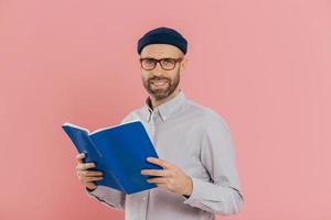 un homme intelligent mal rasé joyeux tient un livre devant lit une histoire passionnante, étudie la littérature scientifique, porte des lunettes et une chemise blanche, se dresse sur fond rose. personnes, lecture, concept de temps libre photo