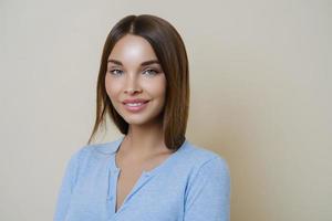 jeune femme brune avec une peau de beauté saine, sourit doucement, montre des dents blanches parfaites, porte un maquillage minimal, vêtue d'un pull décontracté, regarde la caméra et sourit, isolée sur fond beige. photo