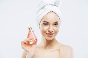 jeune femme à l'air agréable utilise du parfum, aime une nouvelle odeur, se tient ravie à l'intérieur, applique du maquillage, a une peau saine porte une serviette de bain isolée sur fond blanc a un look glamour. odeur agréable photo