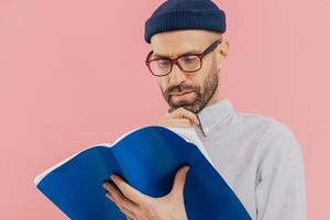 gros plan d'un homme sérieux tient le menton, a du chaume sombre, porte des lunettes pour une bonne vision, porte un livre ouvert, apprend des informations, se tient sur fond rose, impliqué dans la lecture, a un regard intelligent photo