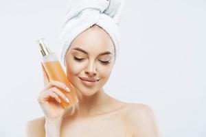 photo horizontale d'une jolie femme avec une peau saine, un maquillage naturel, utilise un vaporisateur de parfum, a une expression agréable, une beauté naturelle, regarde vers le bas, porte une serviette de bain sur la tête, isolée sur fond blanc